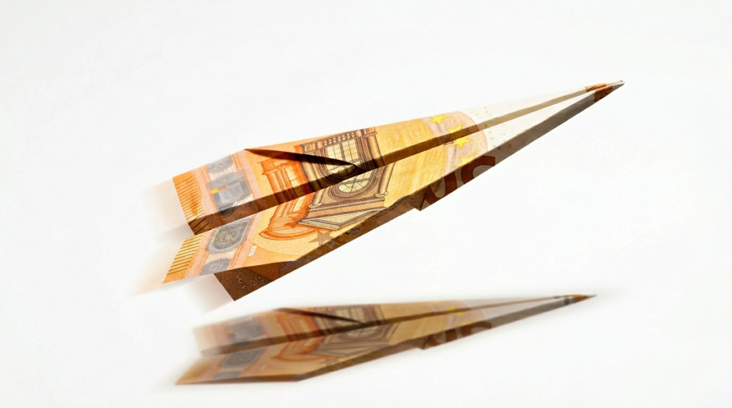 The image features a paper plane expertly folded from a fifty euro banknote, captured mid-flight against a clean white background. The angular folds of the plane display distinct architectural imagery from the note, creating a visual sense of trajectory and optimism, emphasized by the slight upward angle of the nose and the soft shadow beneath.