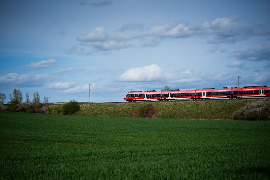 A red and white train travels along a track through lush green fields under a partly cloudy sky. The scene captures the beauty of rural landscapes and train travel. — Credit: Photo by Nico Ruge on Unsplash