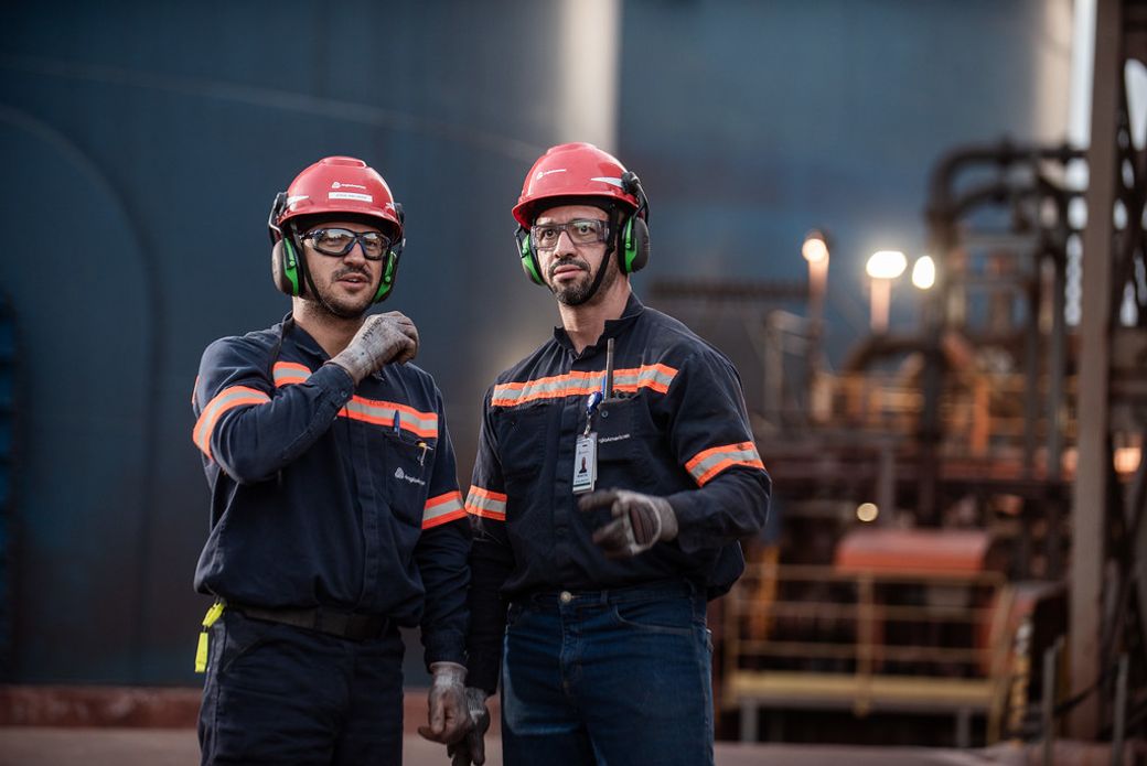Two construction workers wearing helmets and safety gear are standing in an industrial environment, appearing engaged in conversation. The background features machinery and equipment typical of a construction or manufacturing site.