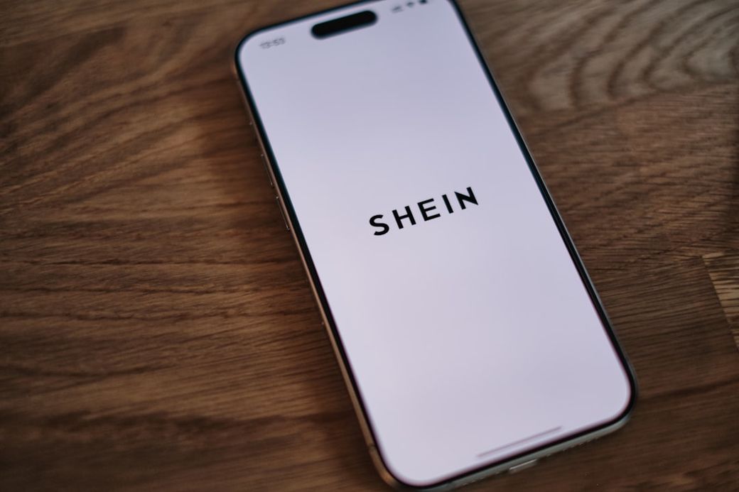 A smartphone displays the logo of Shein, a popular fashion retailer, on its screen. The device is placed on a wooden surface, showcasing a clean and minimalistic aesthetic. — Credit: Photo by appshunter.io on Unsplash
