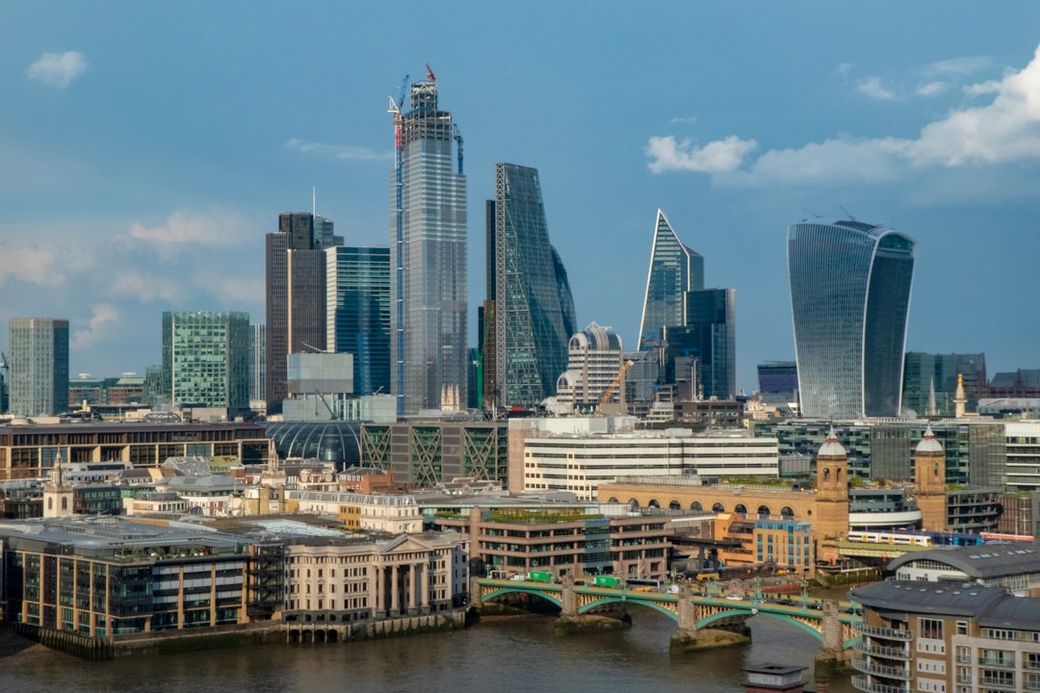 A panoramic view of the London skyline featuring modern skyscrapers, including notable buildings like The Shard and the Walkie Talkie. The River Thames flows through the foreground, with bridges connecting various parts of the city. — Credit: Photo by Nick Fewings on Unsplash