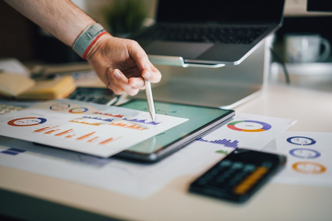 An individual is analyzing data on a tablet while using a stylus. Various charts and graphs printed on papers are displayed on the desk, indicating financial metrics and statistics.