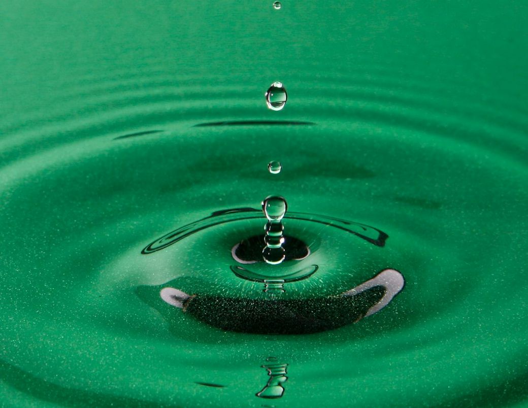 The image features a close-up view of water droplets falling into a green surface, creating ripples in the water. The focus on the water's interaction with the surface highlights the texture and color of the background. — Credit: Photo by Daniele Levis Pelusi on Unsplash