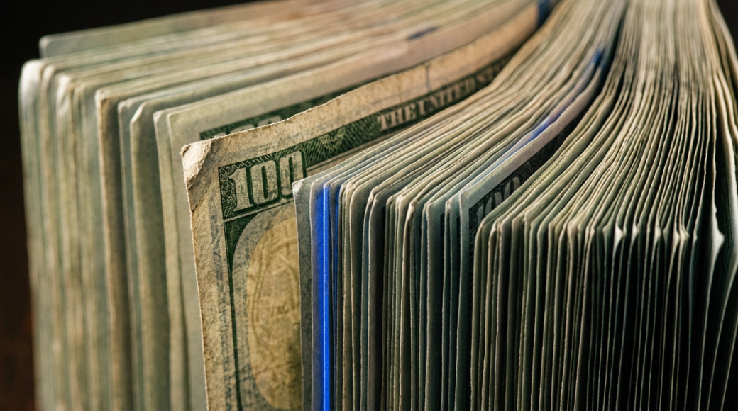 The image showcases a tightly fanned arrangement of U.S. hundred-dollar bills held in a vertical position, emphasizing their textured surfaces and layered edges. Light interacts with the notes, revealing intricate printing details and security features, while a single bill juts slightly forward, its dog-eared corner indicating previous circulation.