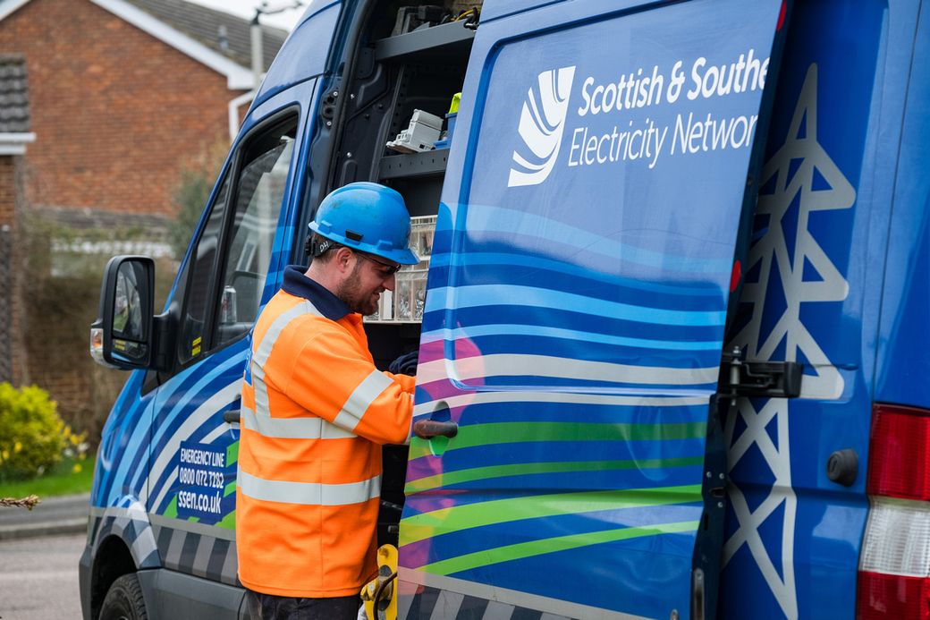 A worker from Scottish & Southern Electricity Network is seen operating a maintenance vehicle. The individual is wearing a blue hard hat and high-visibility orange clothing while examining equipment in the van.