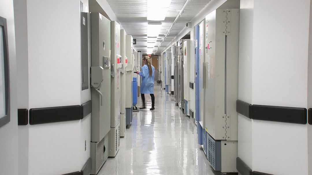 The image depicts a corridor lined with medical freezers and refrigerators in a healthcare facility. A staff member in surgical attire is seen attending to one of the units.