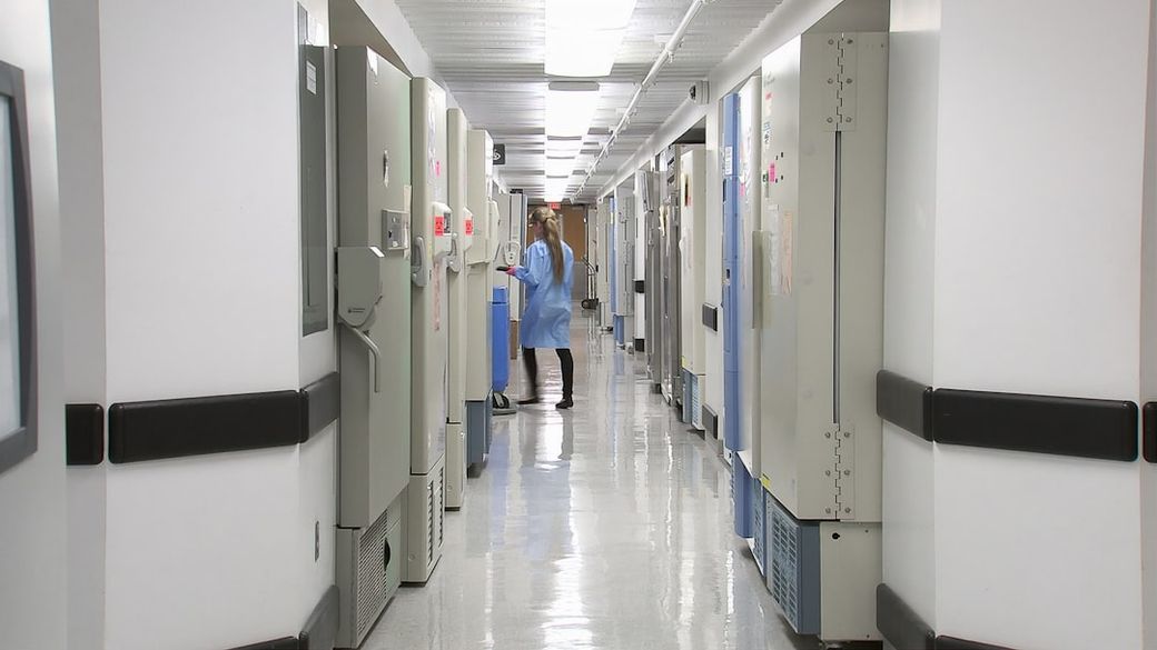 The image depicts a corridor lined with medical freezers and refrigerators in a healthcare facility. A staff member in surgical attire is seen attending to one of the units. — Credit: Photo by CDC on Unsplash