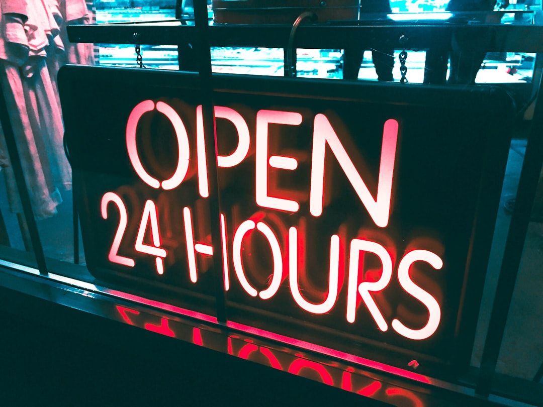 The image features a bright neon sign that reads 'OPEN 24 HOURS', indicating a business that operates continuously. The sign is prominently displayed in a shop window, suggesting accessibility and convenience for customers.