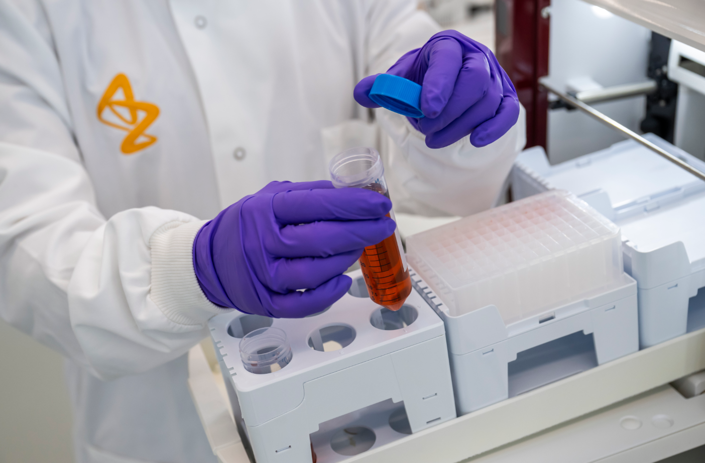 A laboratory technician in a white lab coat and purple gloves is handling a test tube filled with a reddish liquid. The technician is performing an experiment in a modern lab with various laboratory equipment visible in the background.