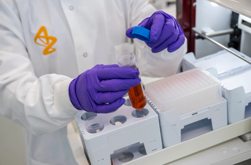 A laboratory technician in a white lab coat and purple gloves is handling a test tube filled with a reddish liquid. The technician is performing an experiment in a modern lab with various laboratory equipment visible in the background. — Credit: Anthony Devlin/Getty Images for AstraZeneca