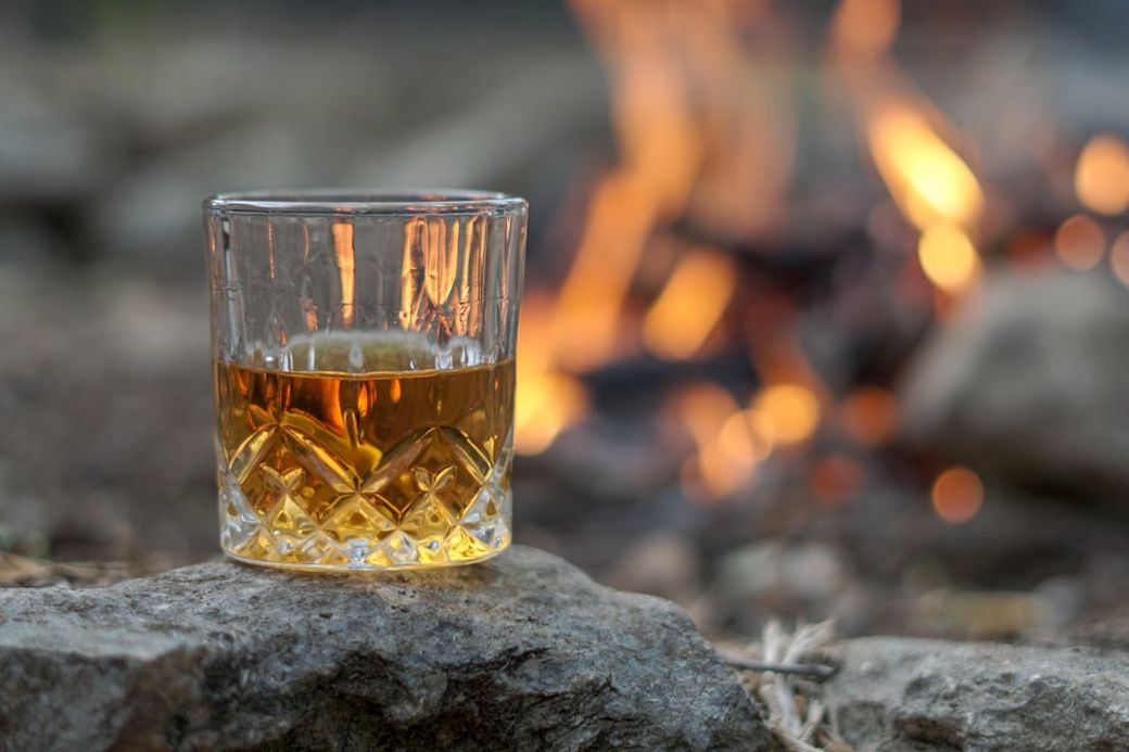 A glass of amber-colored whiskey is placed on a stone against a backdrop of a soft orange and yellow campfire. The glow of the fire contrasts with the clarity of the drink, evoking a warm, inviting atmosphere. — Credit: Photo by Thomas Park on Unsplash