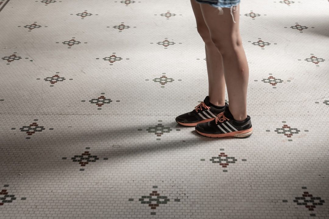 The image depicts a person standing on a tiled floor, showcasing their legs and casual sports shoes. The floor features a patterned design, contributing to the overall aesthetic.
