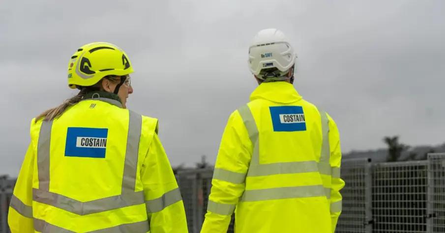 Two construction workers wearing high-visibility jackets and helmets are seen from behind as they walk away from the camera. They are part of a team working on an infrastructure project, with cloudy skies visible in the background.