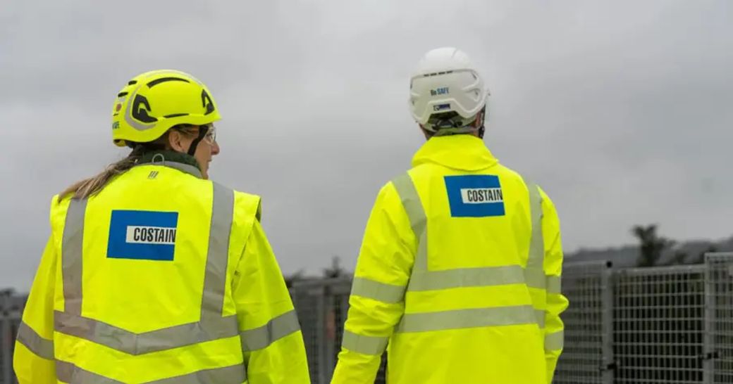 Two construction workers wearing high-visibility jackets and helmets are seen from behind as they walk away from the camera. They are part of a team working on an infrastructure project, with cloudy skies visible in the background.