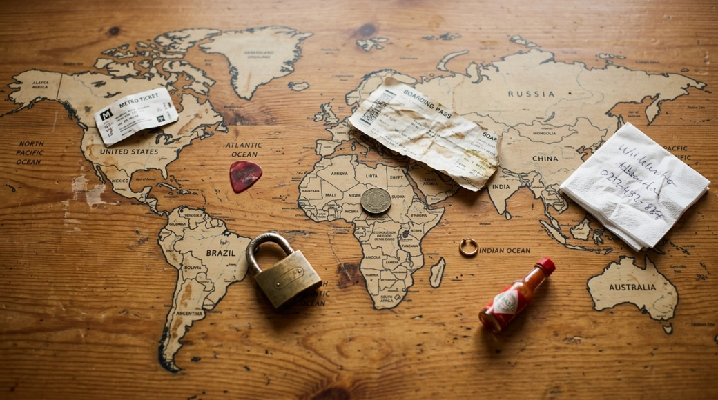 The image presents an abstract overhead view of a world map printed on a wooden table, with various travel-related objects scattered across it. Items like a boarding pass stub, a foreign coin, a luggage padlock, and a miniature bottle of hot sauce lay on different countries, symbolizing the transient experiences of travelers in a hostel setting.