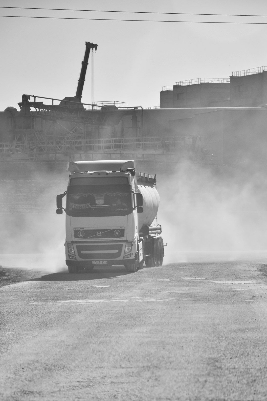 A large industrial truck is driving along a dusty road, surrounded by hazy air and construction machinery in the background. The scene conveys a sense of heavy industry and transportation activity.