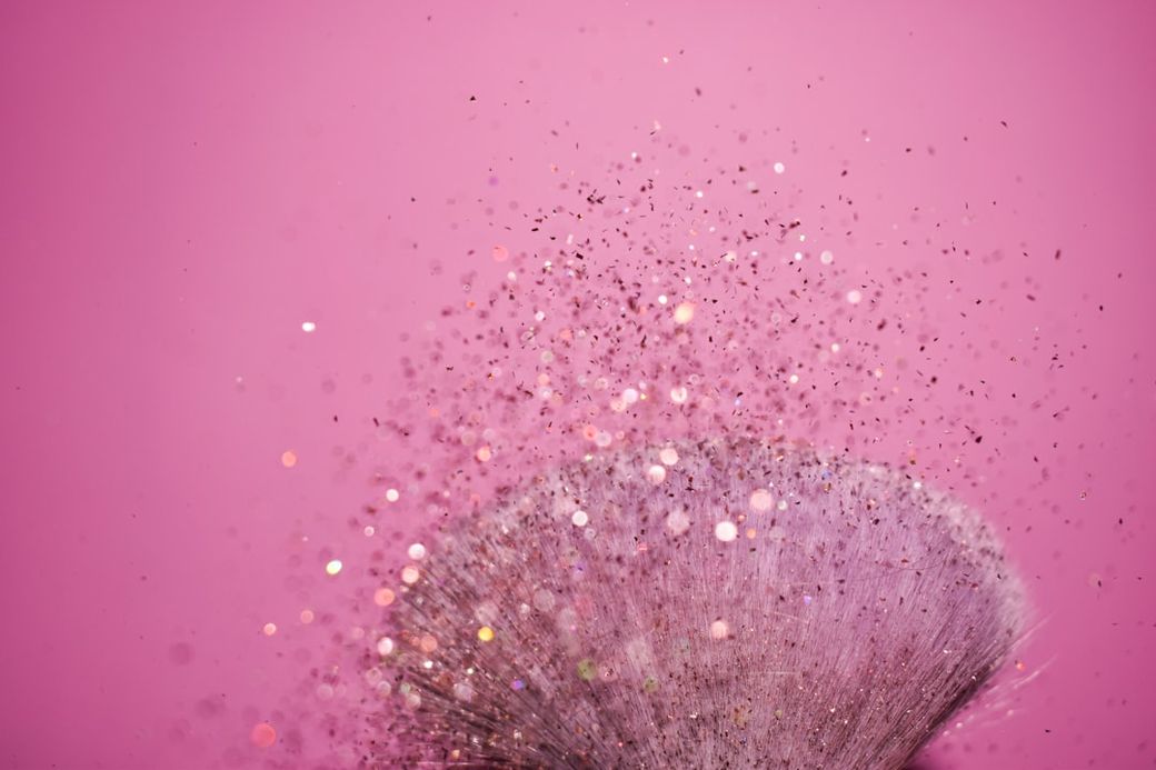 The image features a makeup brush surrounded by shimmering glitter particles against a pink background. The sparkles appear to be in motion, creating a dynamic and vibrant atmosphere. — Credit: Photo by Anna Zyza on Unsplash