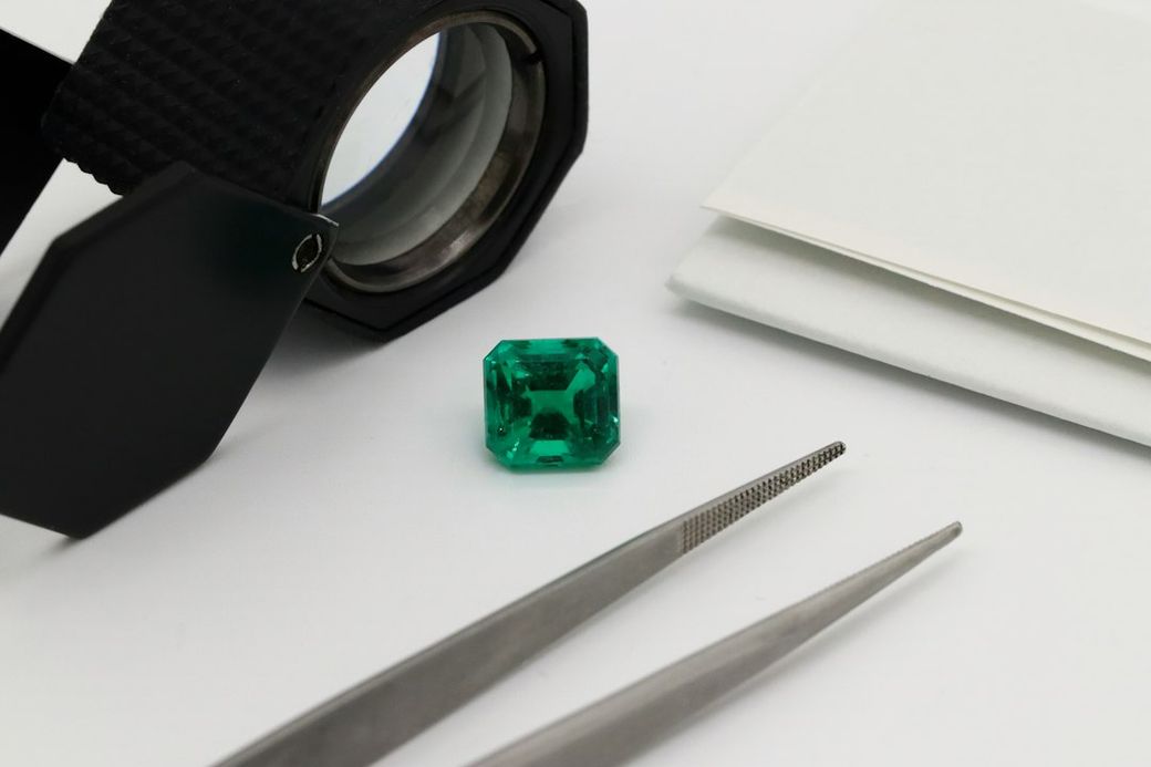The image features a vibrant green emerald positioned on a clean, white surface next to a pair of tweezers and a jeweler's loupe. This setup suggests a context related to gemstone evaluation or jewelry making. — Credit: Photo by Sabrianna on Unsplash