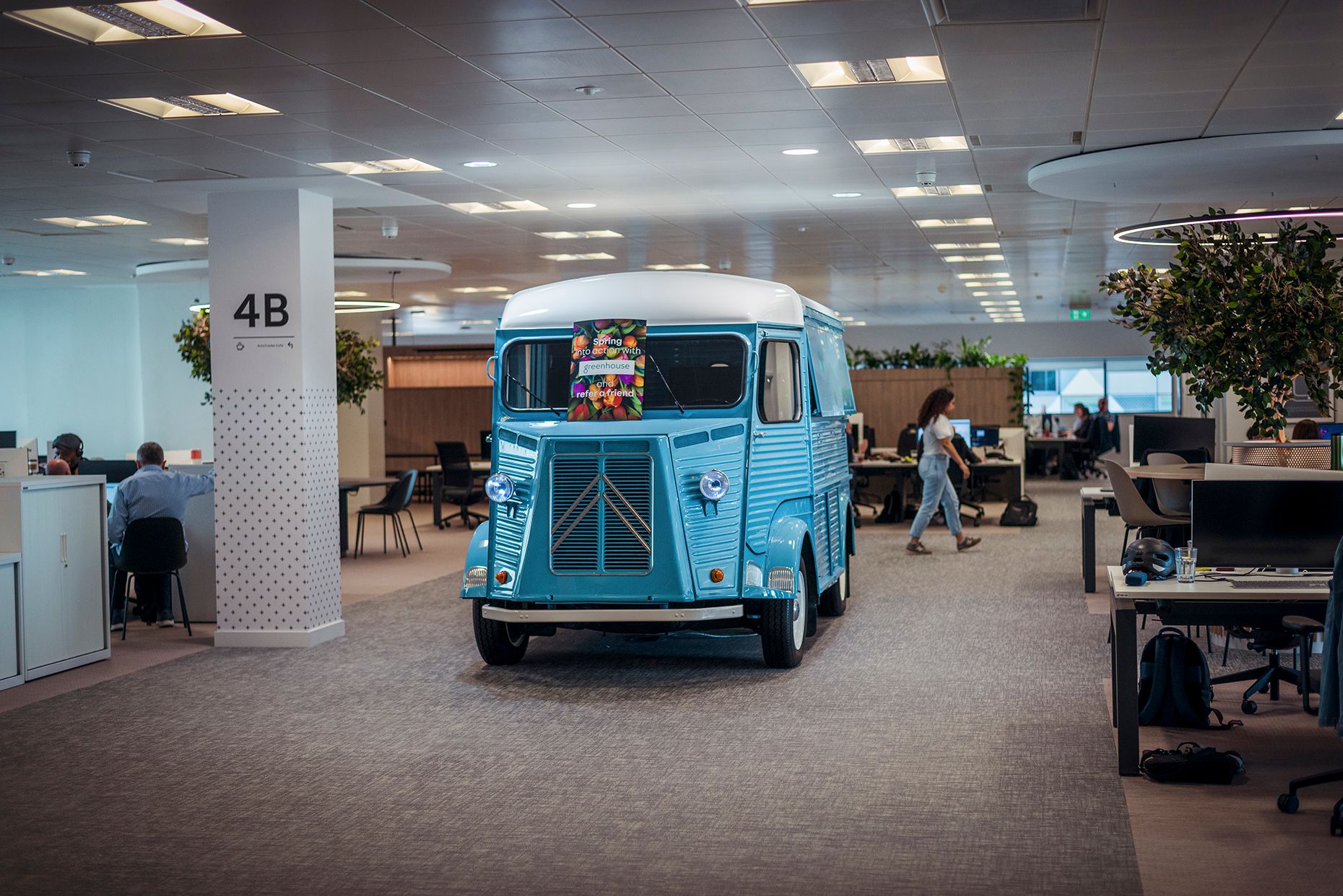 A vintage blue food truck is parked inside a modern office space, surrounded by desks and people working. The truck features decorative items and creates a vibrant atmosphere in the workplace.