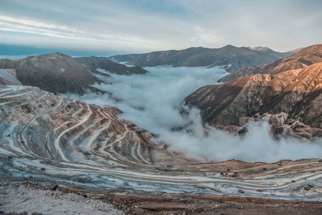 A panoramic view of a mining landscape, showcasing terraced earthworks carved into the mountains. The scene is partially shrouded in fog, with rugged hills rising in the background under a clear sky. — Credit: Photo by omid roshan on Unsplash