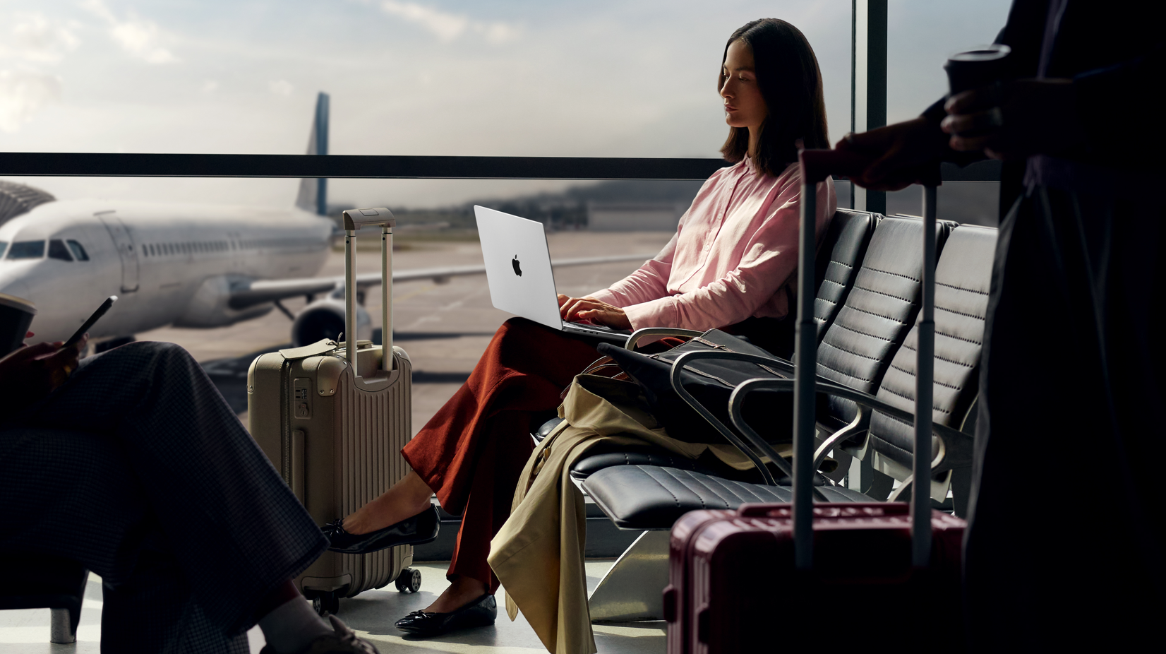 A woman sits at an airport terminal with a laptop open, working while waiting for her flight. In the background, airplanes are visible on the tarmac, suggesting a travel setting.