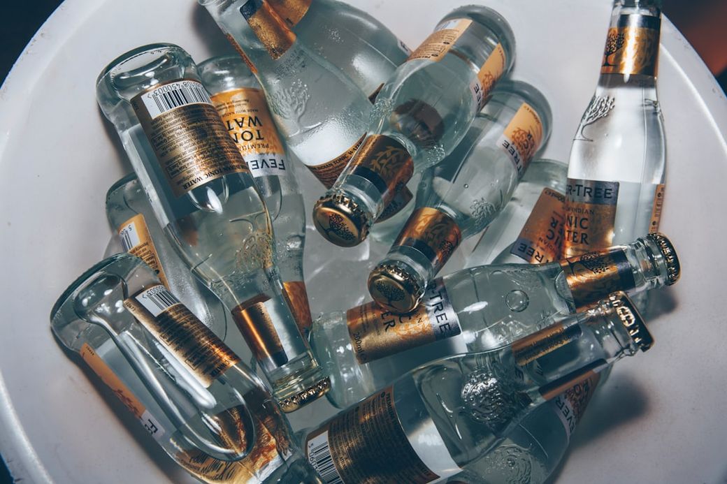 The image shows an arrangement of clear glass bottles filled with a clear liquid, likely tonic water, submerged in ice. The bottles have gold and black labels that indicate branding. — Credit: Photo by Geert Pieters on Unsplash