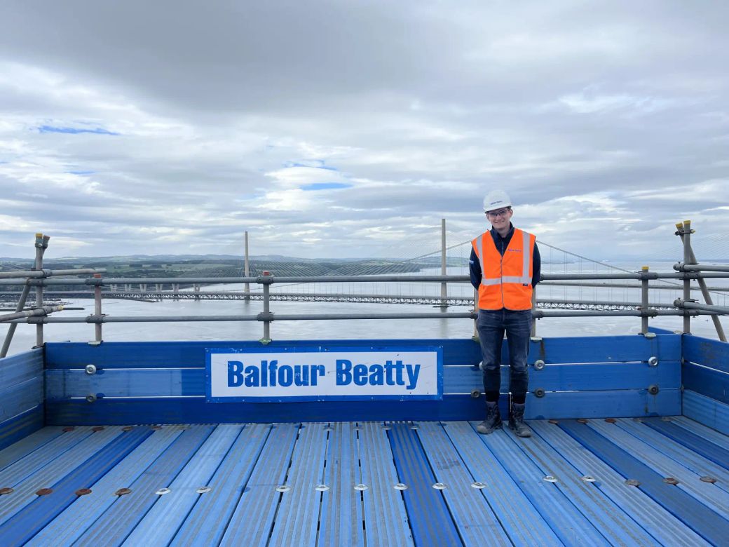 A construction worker stands on a platform overlooking a waterway, with a bridge structure visible in the background. The worker is wearing a hard hat and a high-visibility vest, and the platform is branded with the company name Balfour Beatty.