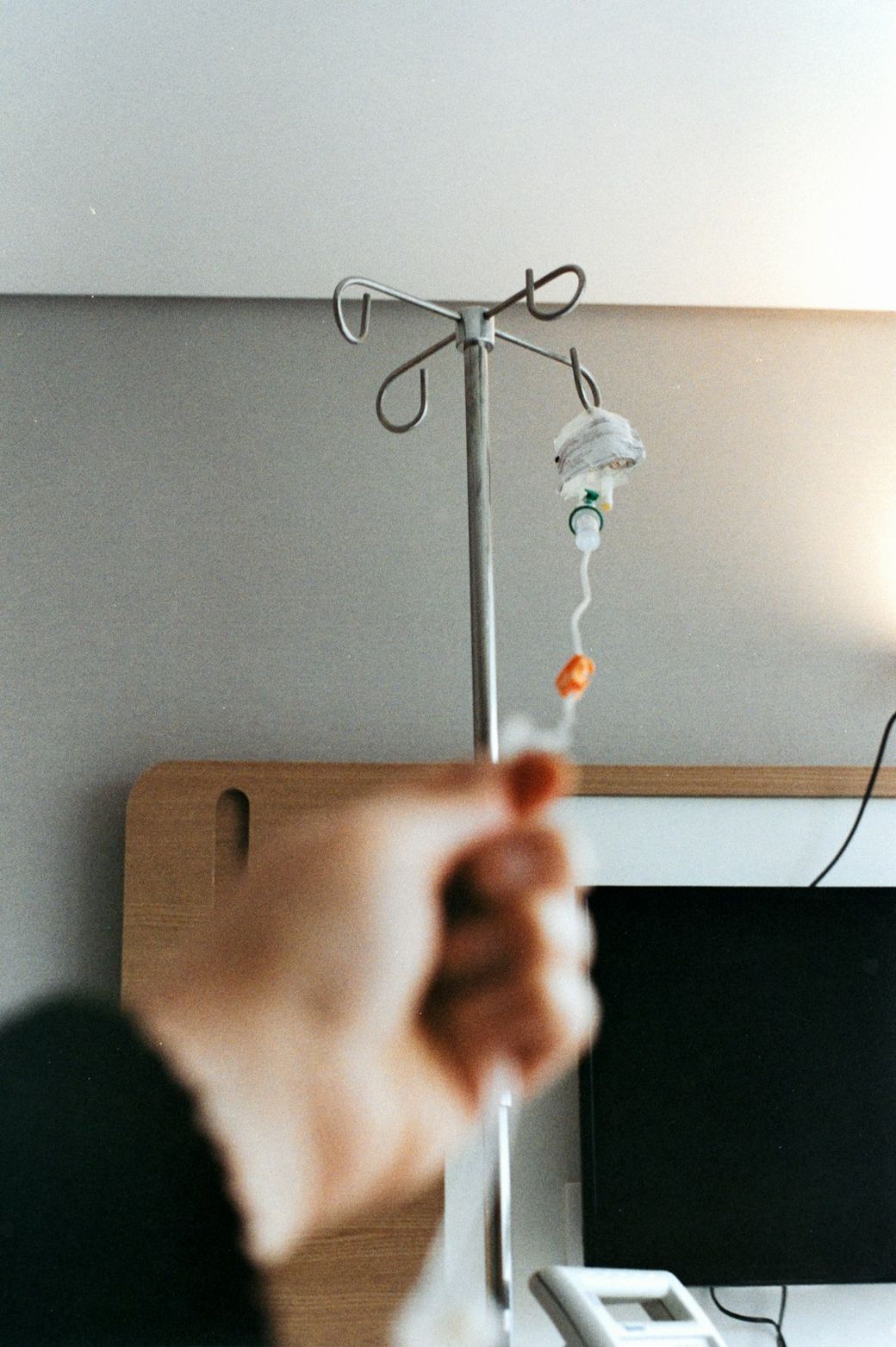 The image depicts a hospital setting with a hand reaching towards an intravenous (IV) drip stand. The background includes medical equipment and a shaded wall, suggesting a patient care area. — Credit: Photo by Camila Mofsovich on Unsplash