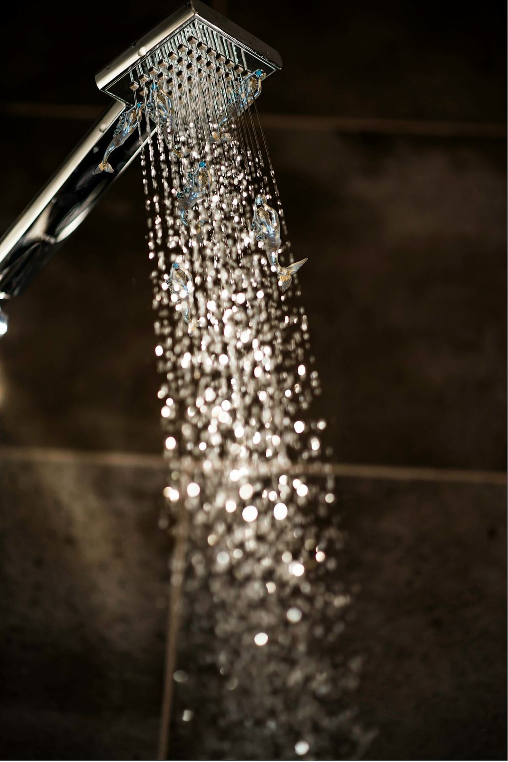 A close-up image of a modern showerhead with water cascading down in droplets. The background features dark tiles, highlighting the sleek design of the shower fixture. — Credit: Photo by Ginger Hendee on Unsplash