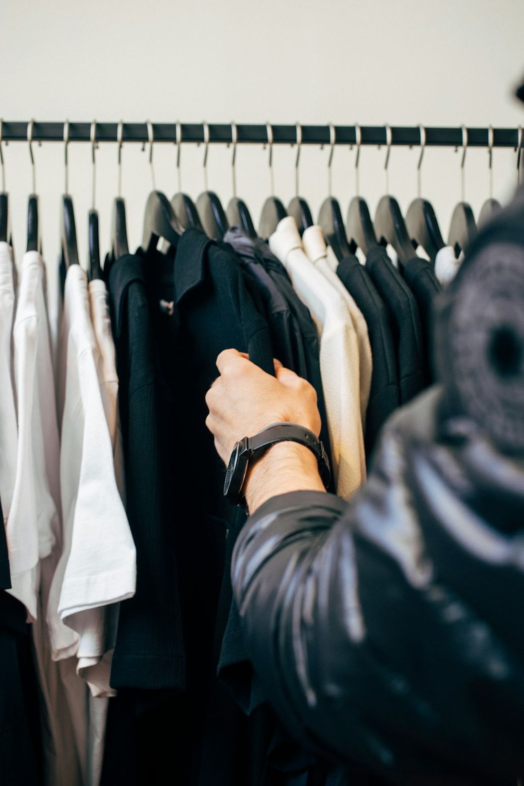 A close-up image of a person selecting a t-shirt from a clothing rack. The rack displays a variety of t-shirts in different colors, including black, white, and beige. — Credit: Photo by charlesdeluvio on Unsplash