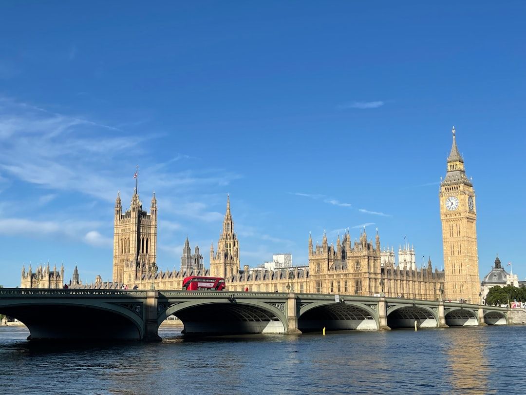 The image showcases the iconic Palace of Westminster in London, featuring the Houses of Parliament and the Elizabeth Tower, commonly known as Big Ben. A red double-decker bus crosses the bridge over the River Thames, under a clear blue sky.