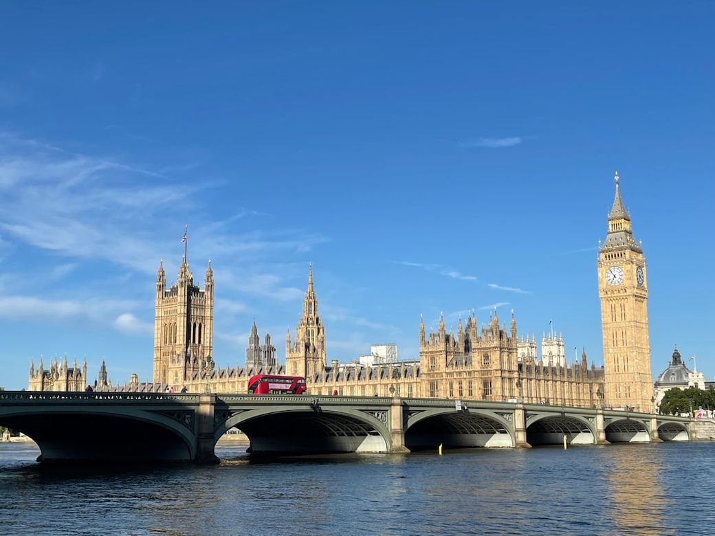 The image showcases the iconic Palace of Westminster in London, featuring the Houses of Parliament and the Elizabeth Tower, commonly known as Big Ben. A red double-decker bus crosses the bridge over the River Thames, under a clear blue sky. — Credit: Photo by N R on Unsplash