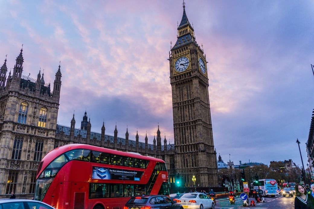 The image depicts a red double-decker bus in motion near the iconic Big Ben clock tower in London, set against a colorful sky at dusk. The scene captures a busy urban atmosphere with both vehicles and pedestrians. — Credit: Photo by Lucas Davies on Unsplash