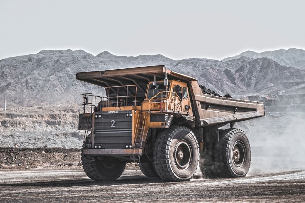 A large heavy-duty dump truck drives through a mining site, with a mountainous landscape in the background. Dust is kicked up from the ground, indicating active excavation. — Credit: Photo by omid roshan on Unsplash