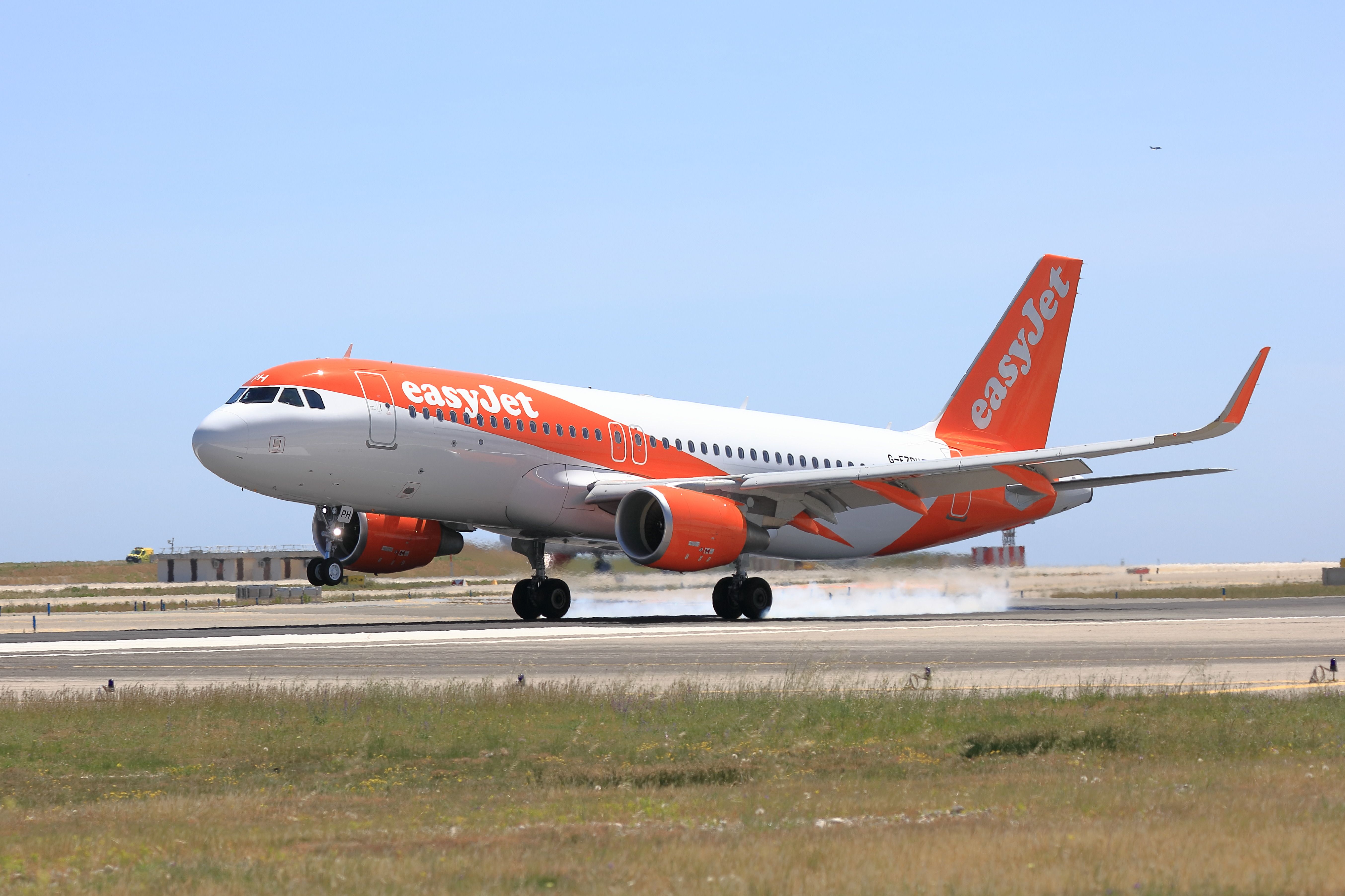An easyJet airplane is shown taking off from a runway, with its wheels just above the ground. The aircraft is prominently displaying the easyJet branding with a clear blue sky in the background.