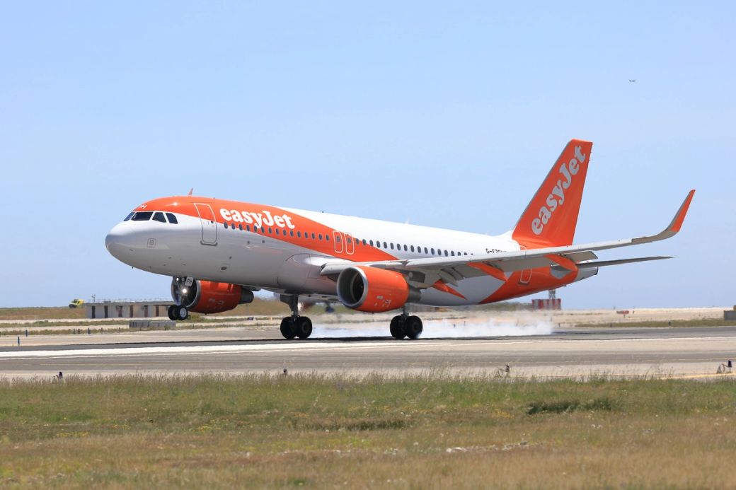 An easyJet airplane is shown taking off from a runway, with its wheels just above the ground. The aircraft is prominently displaying the easyJet branding with a clear blue sky in the background.
