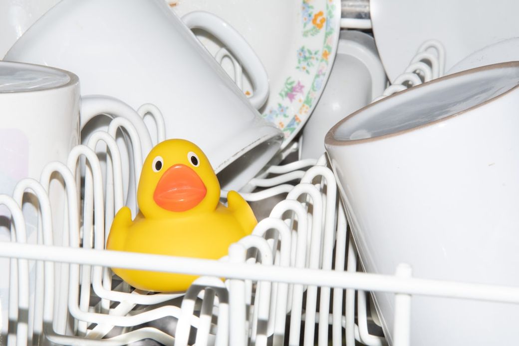 The image shows a yellow rubber duck amidst several white mugs inside a dishwasher. The duck is positioned prominently, contrasting with the surrounding kitchenware. — Credit: Photo by Pavol Tančibok on Unsplash