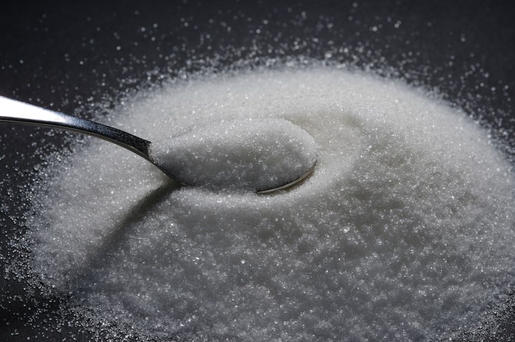 The image features a close-up shot of a spoonful of granulated sugar placed on a mound of sugar. The focus is on the fine texture of the sugar grains against a dark background. — Credit: Photo by Immo Wegmann on Unsplash