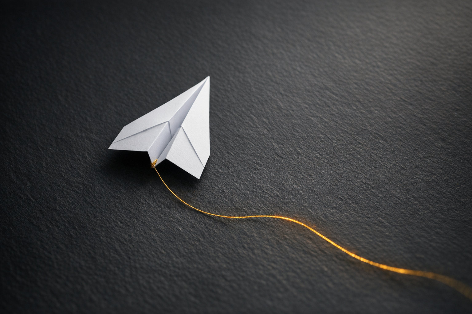 The image features a paper airplane crafted from white paper, positioned on a dark textured surface. A thin, golden thread is attached to the airplane, suggesting an artistic or conceptual theme related to flight or communication.