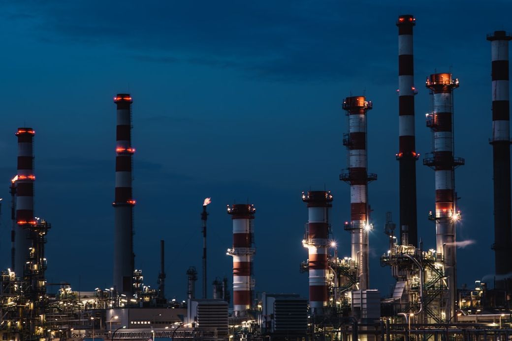 The image depicts a nighttime industrial scene featuring several tall smokestacks with red and white stripes illuminated against a dark blue sky. Water vapor and lights are visible, suggesting activity at a manufacturing or energy facility. — Credit: Photo by Maksym Kaharlytskyi on Unsplash