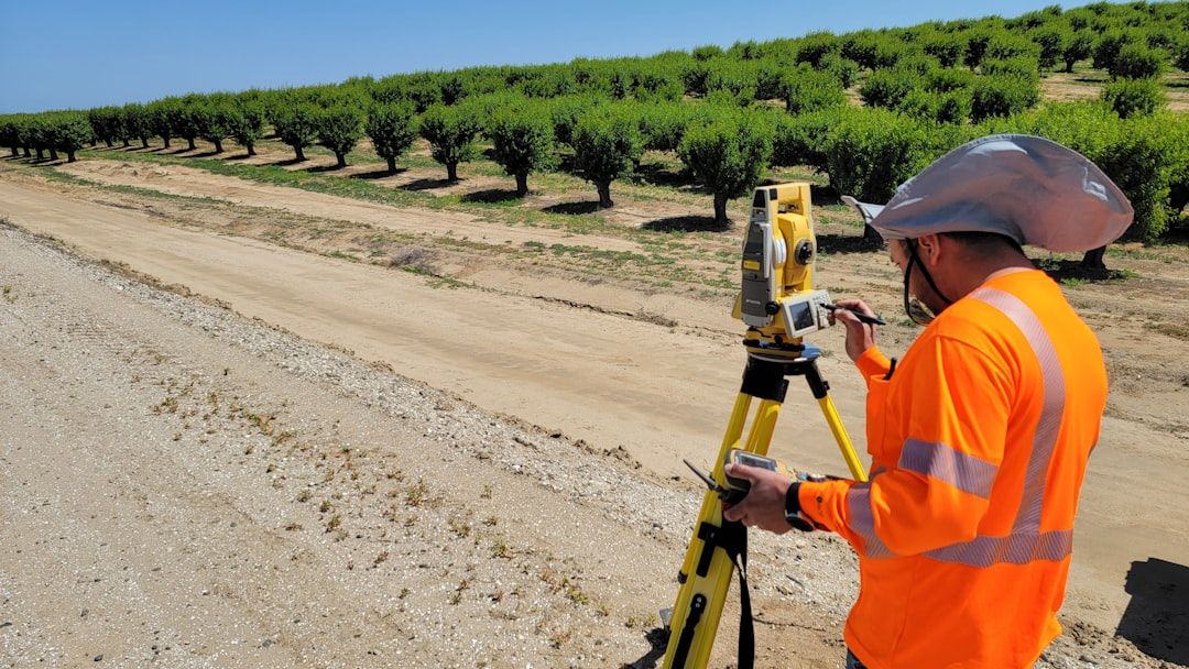 A land surveyor is using a surveying instrument on a dirt road next to a row of trees in an agricultural field. The surveyor is wearing a high-visibility orange shirt and a sun hat, indicating outdoor work in a sunny environment.