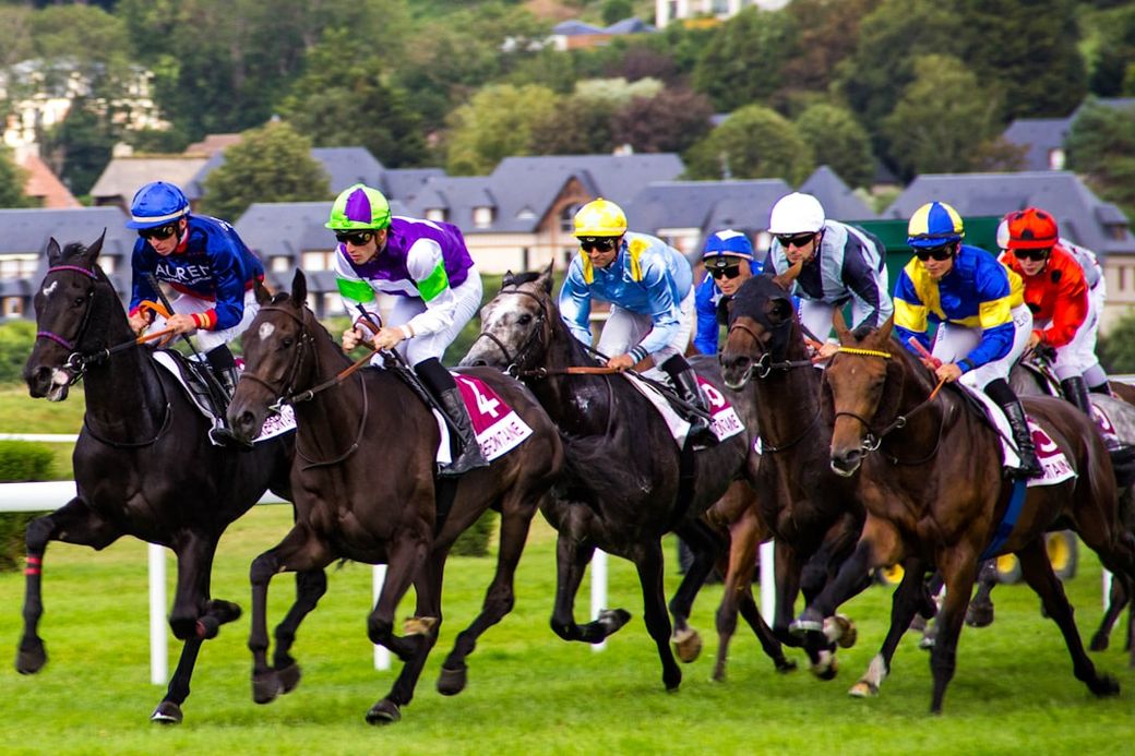 A group of jockeys racing on horseback during a horse racing event. The race features multiple horses competing on a green racetrack, with spectators and a hillside backdrop. — Credit: Photo by Philippe Oursel on Unsplash