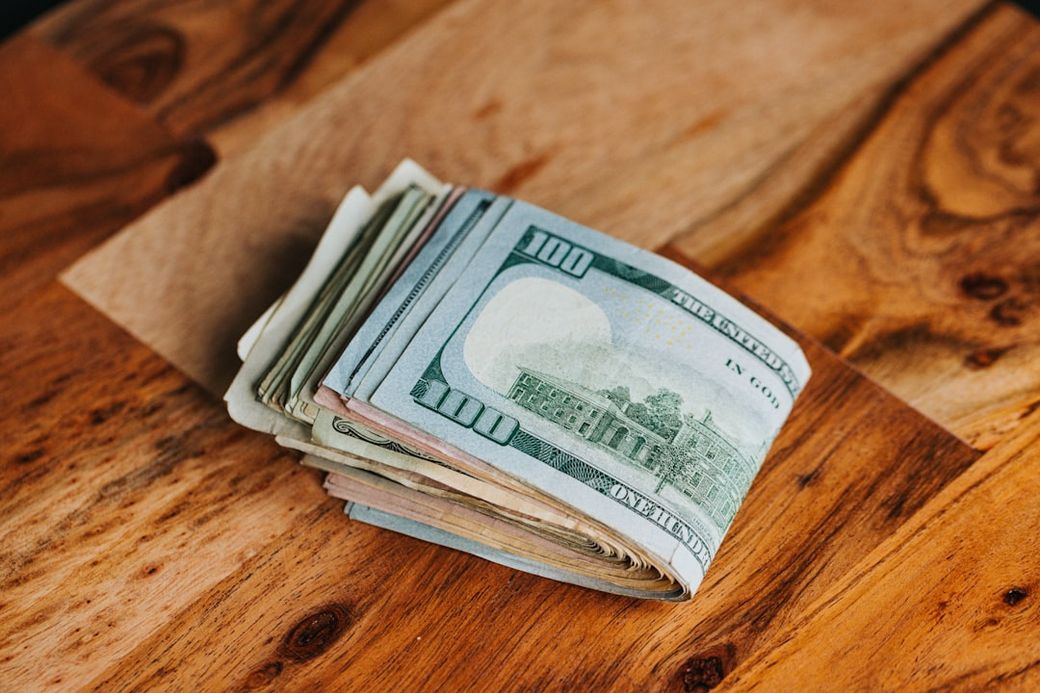 A stack of U.S. dollar bills placed on a wooden surface, showcasing various denominations. The image focuses on a prominent stack of hundred-dollar bills, emphasizing financial currency. — Credit: Photo by Nathan Dumlao on Unsplash