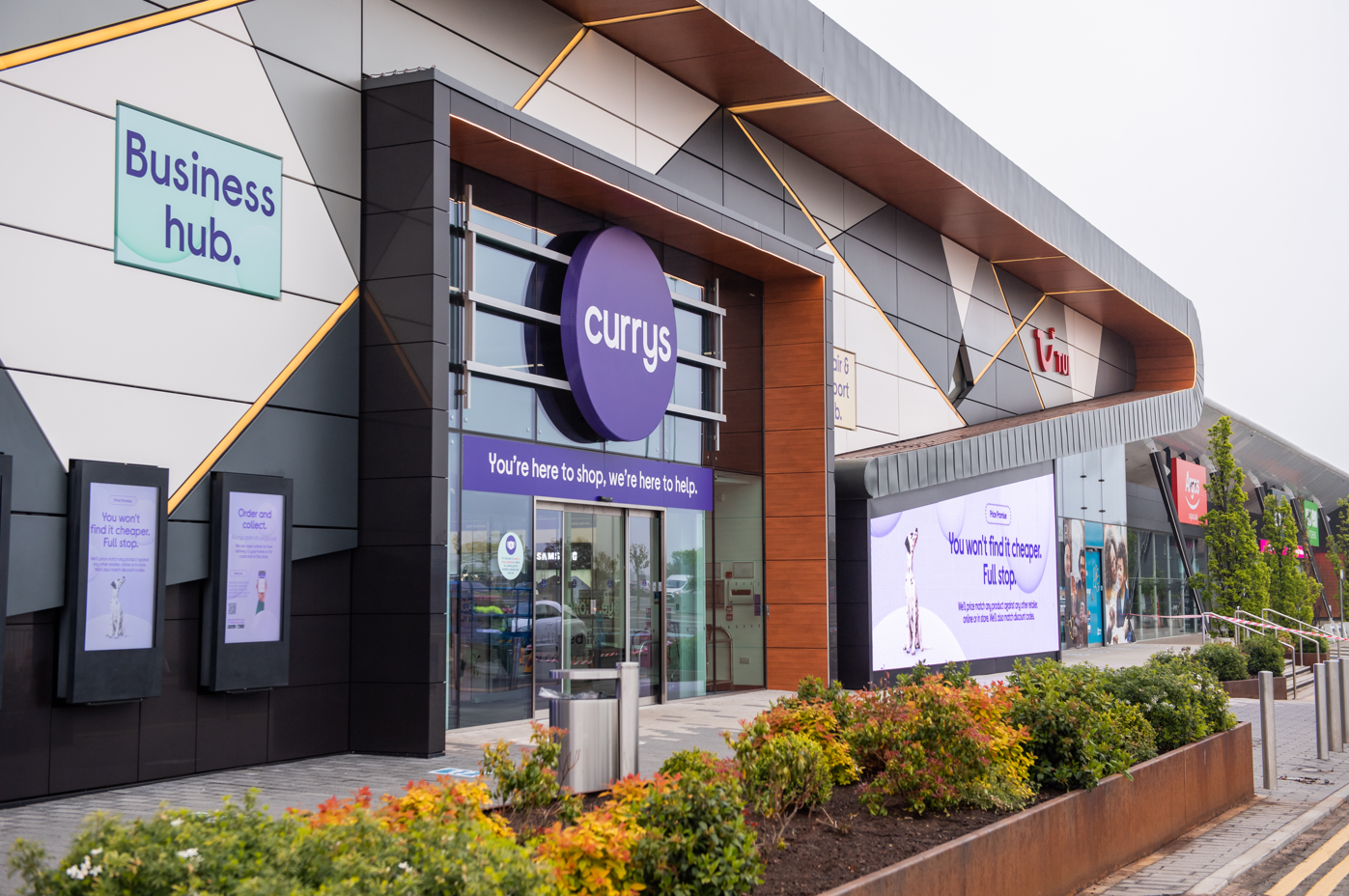 The image shows a retail storefront featuring the 'Currys' brand, located in a modern shopping center. Surrounding the entrance are digital displays with various promotional messages.