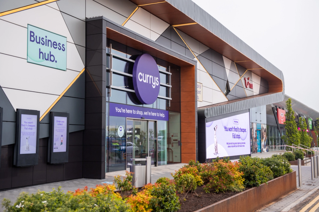 The image shows a retail storefront featuring the 'Currys' brand, located in a modern shopping center. Surrounding the entrance are digital displays with various promotional messages.