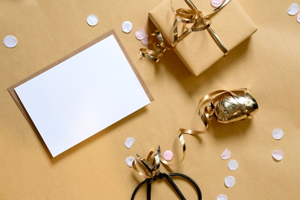 The image features a beautifully wrapped gift box with a shiny ribbon, accompanied by a blank greeting card and scattered confetti on a plain background. It conveys a festive atmosphere suitable for celebrations such as birthdays or holidays. — Credit: Photo by Jess Bailey on Unsplash