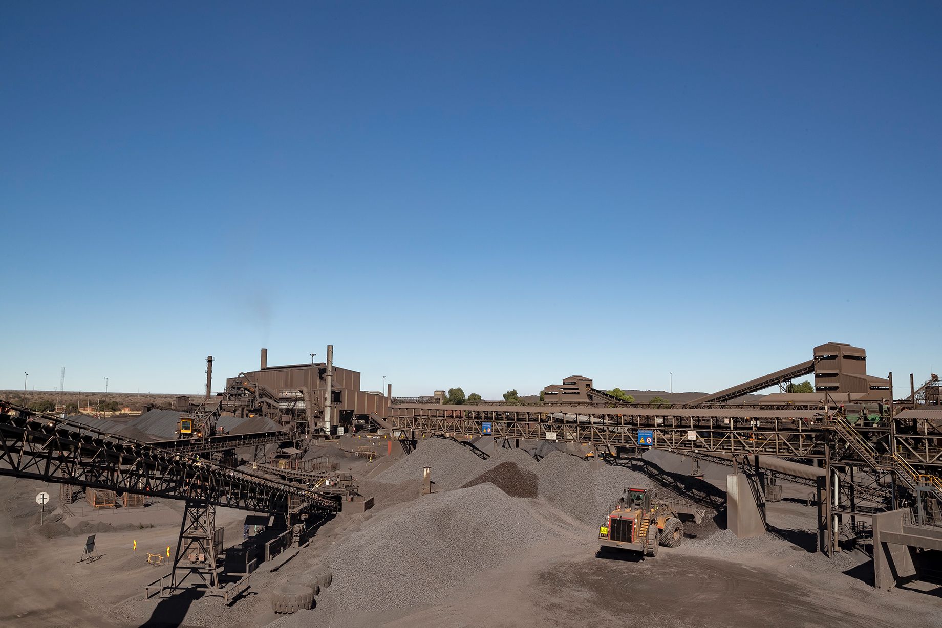 The image depicts a large coal mining facility featuring extensive machinery and infrastructure. Piles of coal and mining equipment are visible under a clear blue sky, illustrating the scale of industrial operations.