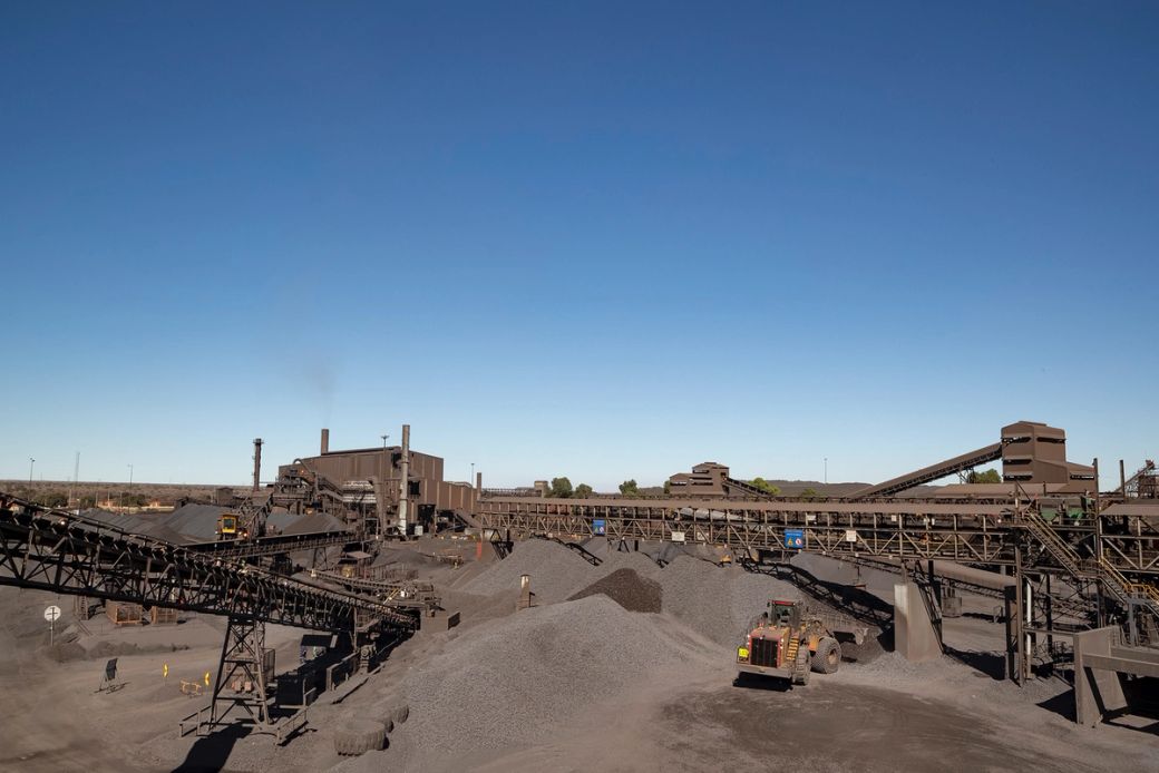 The image depicts a large coal mining facility featuring extensive machinery and infrastructure. Piles of coal and mining equipment are visible under a clear blue sky, illustrating the scale of industrial operations.