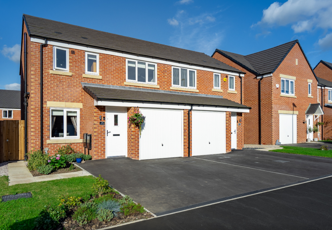 The image showcases a row of modern brick houses with double garages and well-maintained gardens. The sky is clear and blue, highlighting the residential nature of the area.