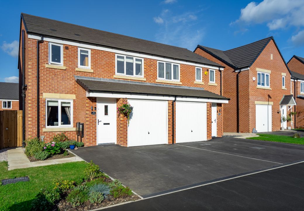 The image showcases a row of modern brick houses with double garages and well-maintained gardens. The sky is clear and blue, highlighting the residential nature of the area.